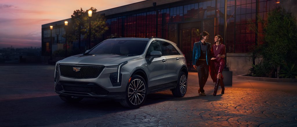 A silver Cadillac XT4 sits outside a modern building at dusk as a stylish couple walks by.