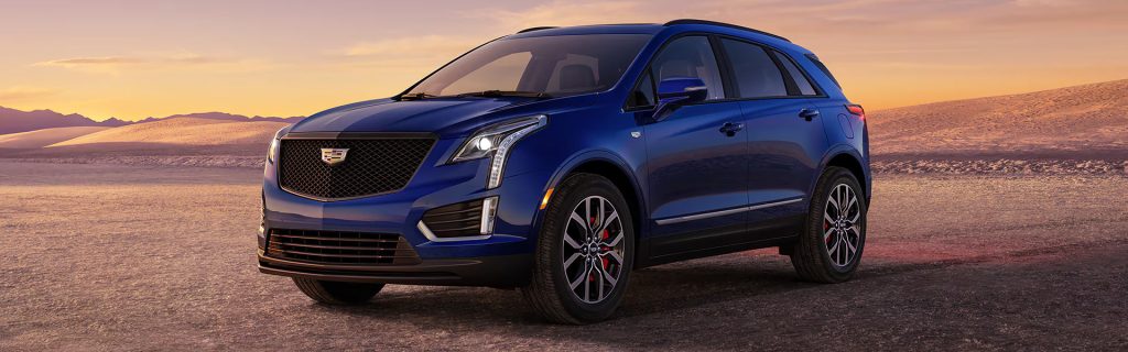 Blue Cadillac SUV parked on a desert landscape at sunset, with mountains in the background and warm orange sky.