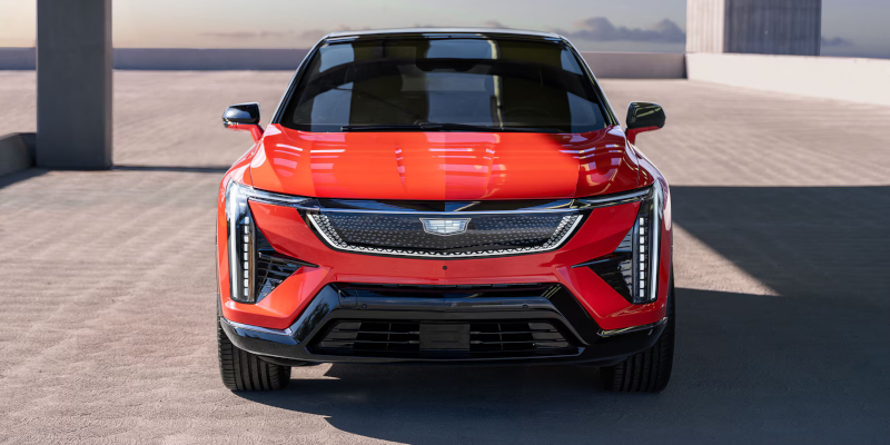 A front view of a red modern electric SUV parked in an open outdoor area, featuring sleek LED headlights, a black roof, and a futuristic grille design.