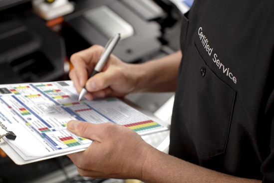 A service technician wearing a "Certified Service" shirt fills out a vehicle inspection checklist on a clipboard.