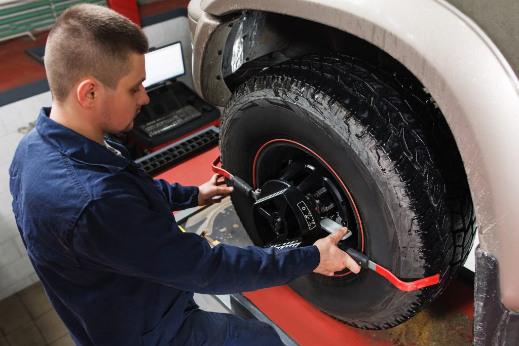 A mechanic in blue coveralls attaches a wheel alignment clamp to a truck's front tire.
